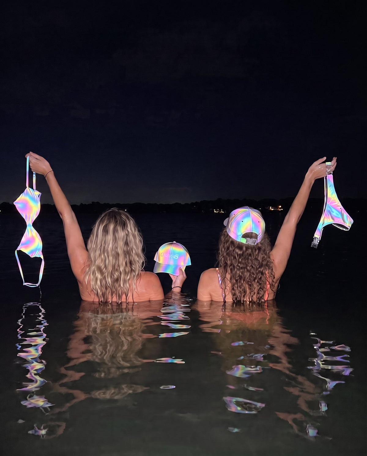 Two people in the water at night holding up the Clickinis iridescent bikini top, bottom and cap, reflecting the flash